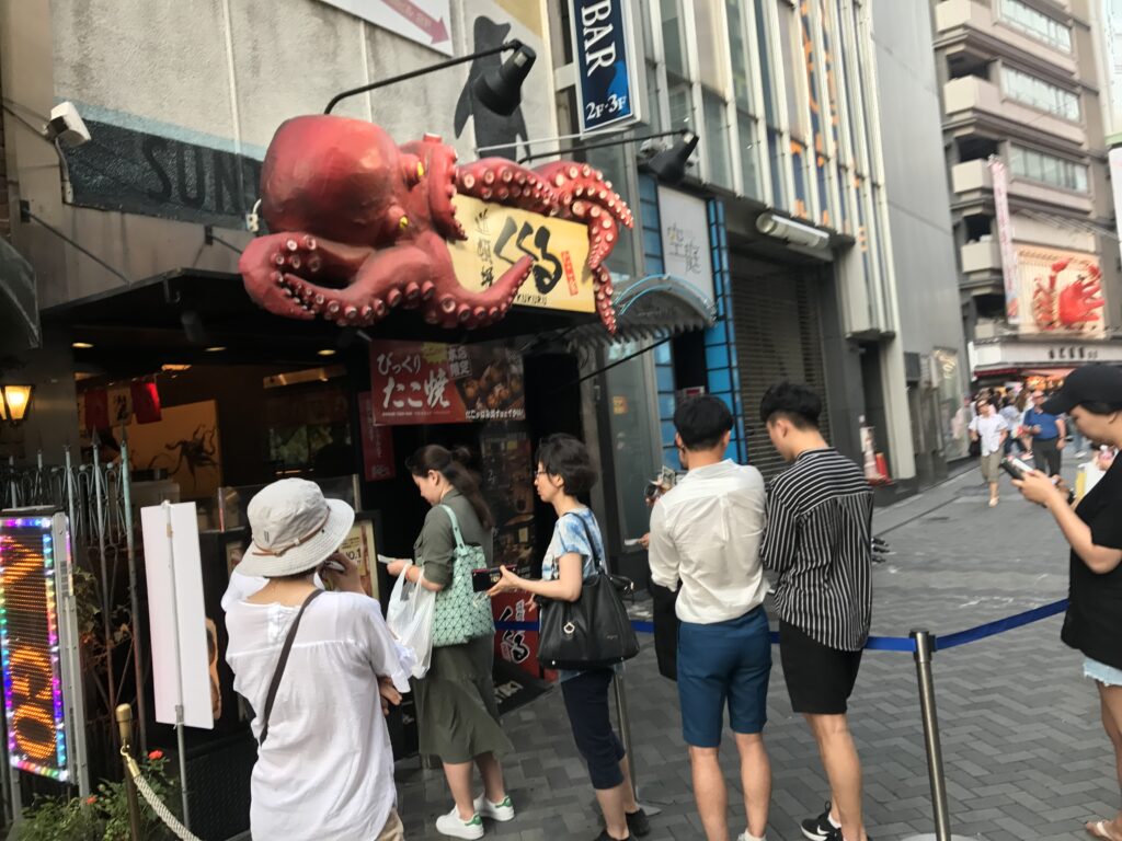 大阪・道頓堀の人気たこ焼き店「くくる」に並ぶ観光客たち。巨大なタコのオブジェが目を引く店舗外観。