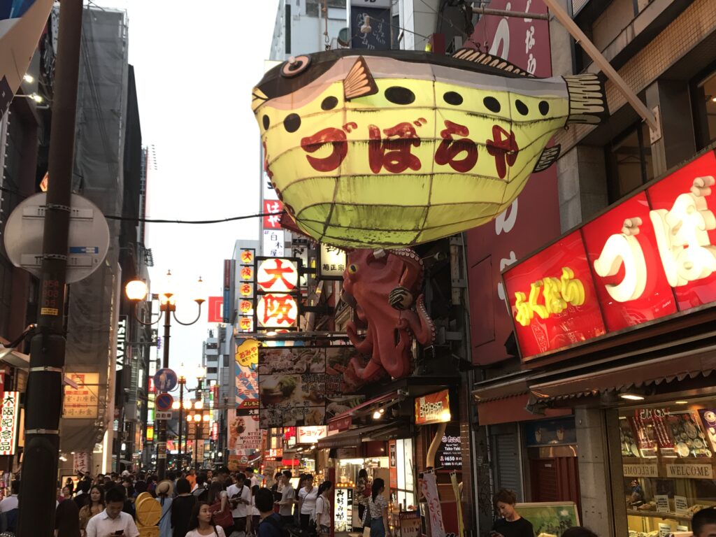 道頓堀の繁華街に掲げられた「づぼらや」の巨大なフグ提灯とタコの看板。夕暮れ時のにぎわいがあふれる通りの様子。