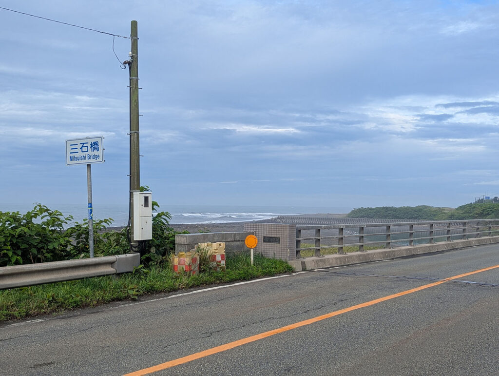 三石橋の道路と欄干、橋名標識とプレート。背後には太平洋の波が広がる。