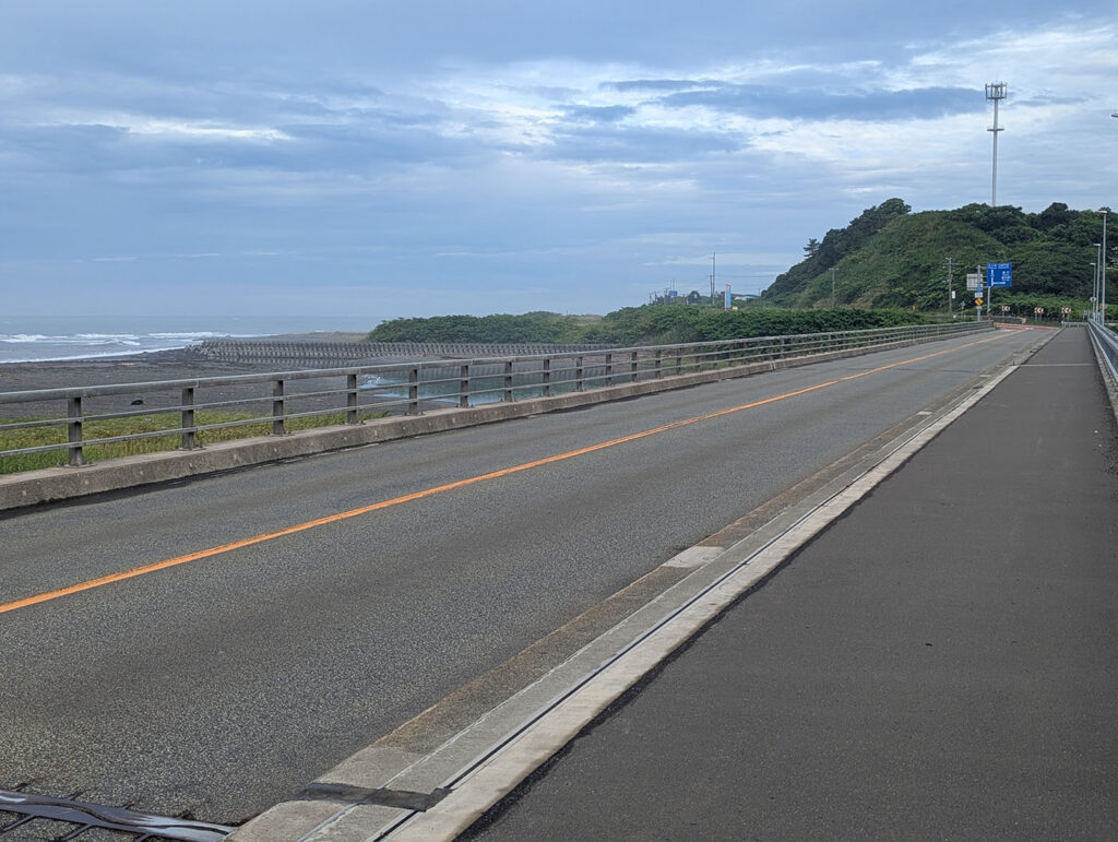 三石橋の車道と歩道、橋の先に緑の丘、右奥に道路標識と照明塔が見える風景。