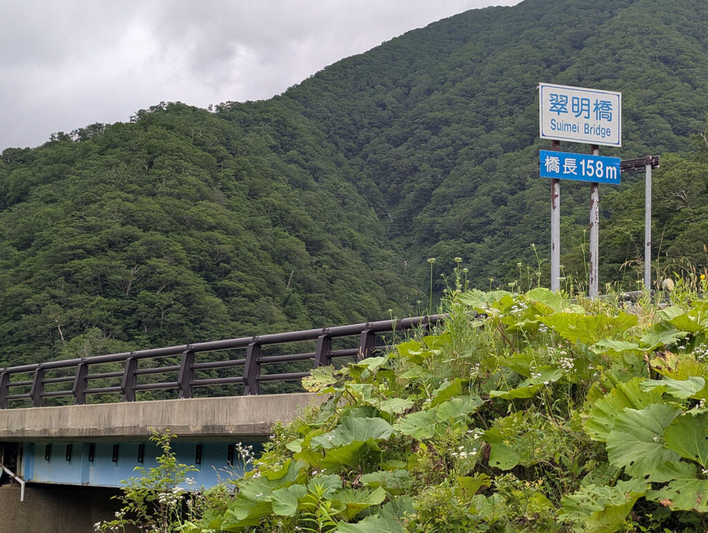 北海道浦河町の翠明橋(Suimei Bridge)の標識と橋長158メートルの案内板が立つ青い橋。山の緑に囲まれた静かな風景の中に位置する。