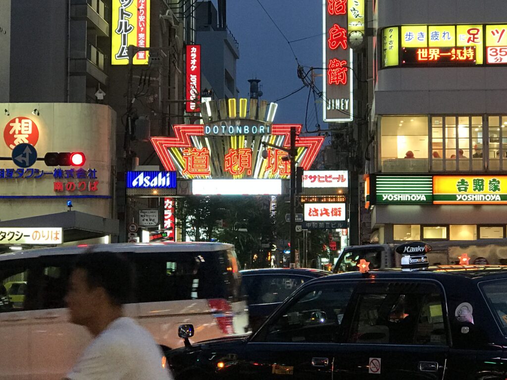 大阪・道頓堀の象徴的なネオンサイン「道頓堀」ゲート。夕暮れ時の交差点にはタクシーや歩行者が行き交い、街のにぎわいが広がる。