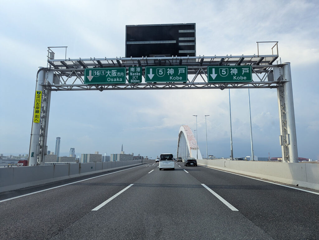 高速道路上、大阪市内方面と神戸方面への分岐案内標識。正面には赤い大きなアーチ橋が見えている。