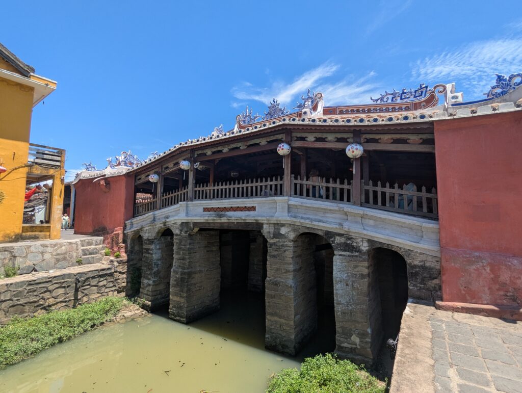青空の下、瓦屋根と精緻な龍の装飾が施された木造の屋根付き橋。石造りの橋脚の上に、赤壁と格子柵が連なる歴史的建造物が立つ。