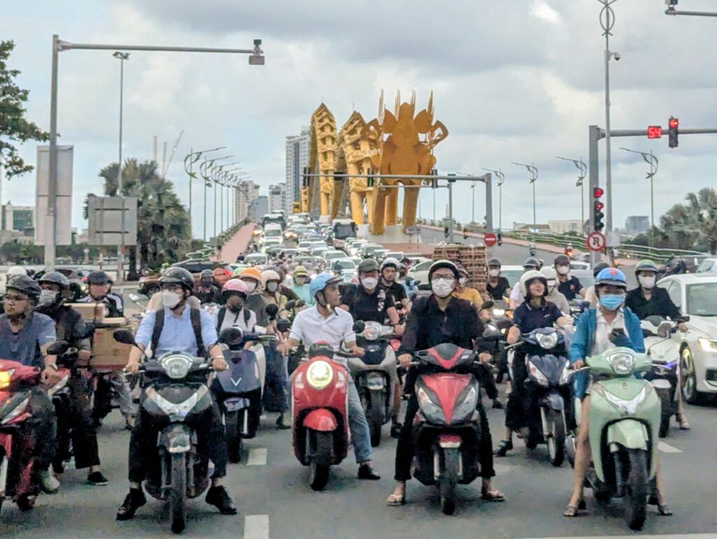 ベトナム・ダナンのドラゴンブリッジ前で信号待ちをする大量のバイクとライダー。黄金の龍の彫刻が奥にそびえ、まるで吐き出された騎兵隊のように見える光景」