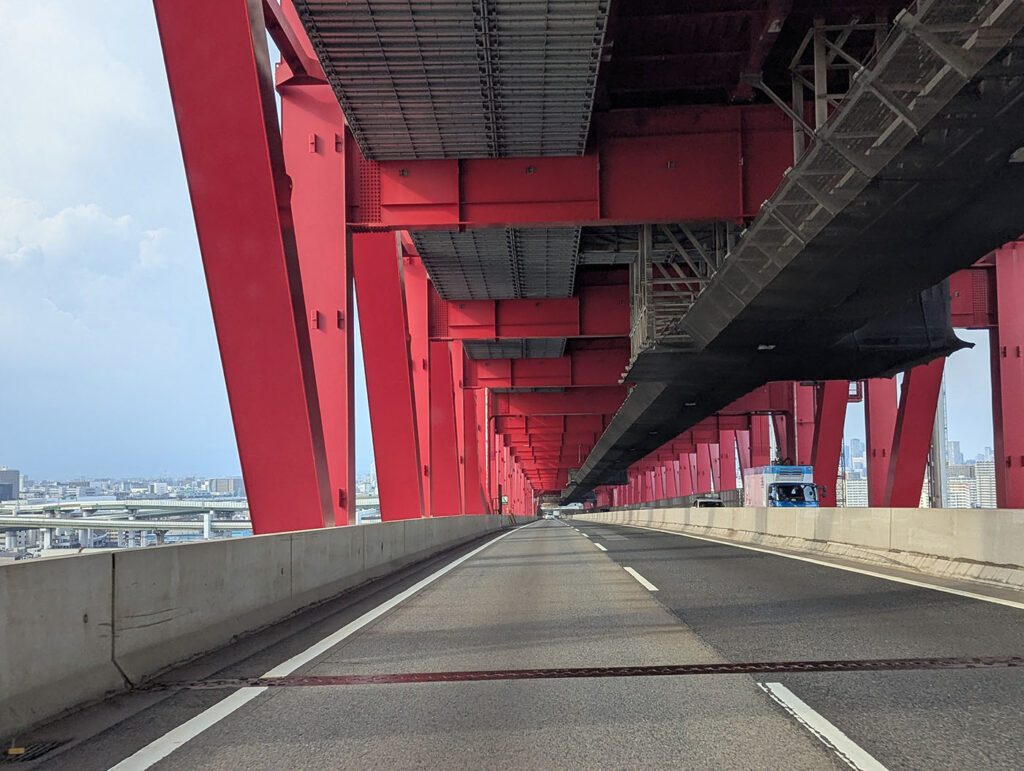 真っ赤な鉄骨に囲まれた高速道路の橋梁内部。頭上に複雑な鉄骨構造が続き、奥へと伸びる道が力強さを感じさせる。
