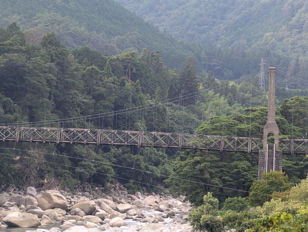 長野県南木曽町にかかる桃介橋。木製トラスと鉄線で構成された吊り橋が、緑深い山あいの谷を横切る。下には岩が転がる木曽川の流れが見える。