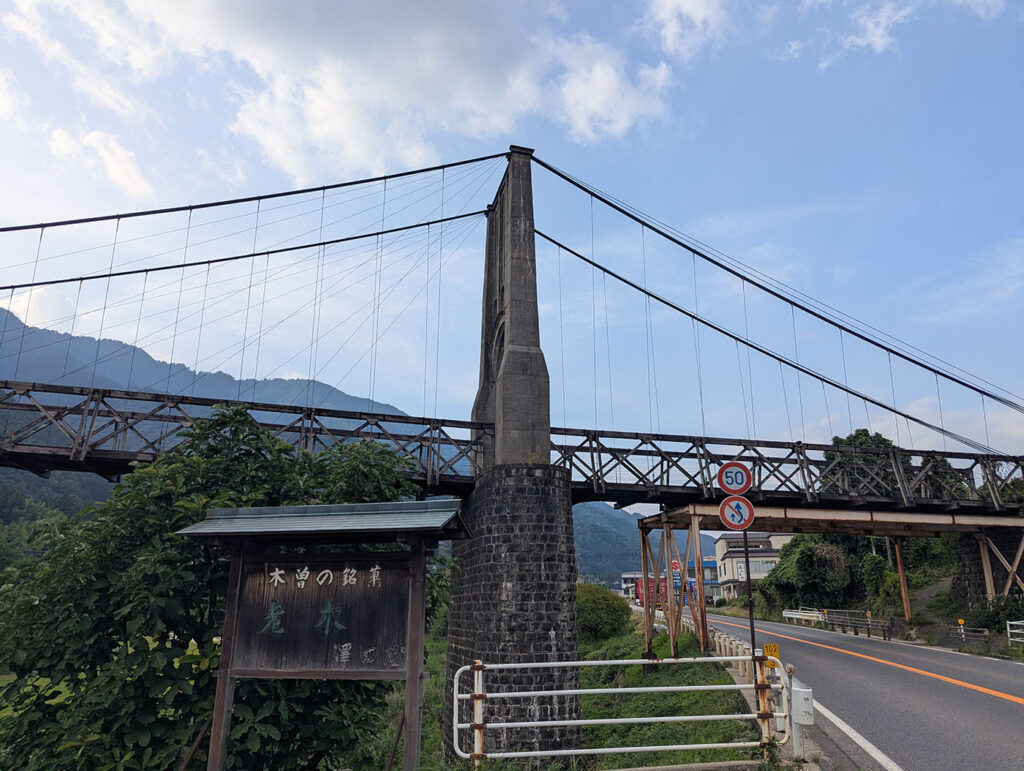 国道沿いから見上げた桃介橋。石造りの橋脚と木製トラスが力強く立ち、青空にワイヤーが美しく伸びている。