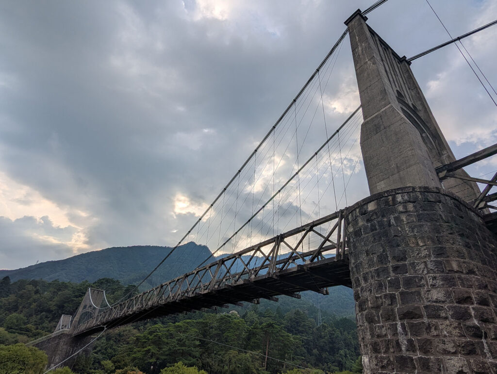 曇り空の下、石造りの橋脚とワイヤーがそびえる桃介橋。背後に山がそびえ、夕方の光が雲間から差し込む。