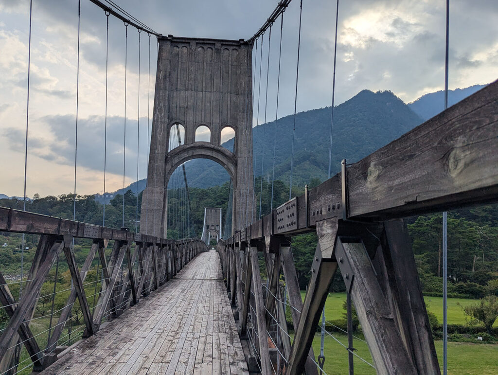 桃介橋の木製デッキを進む視点。アーチ状の主塔が正面に並び、ワイヤーが山の稜線へと続いている。