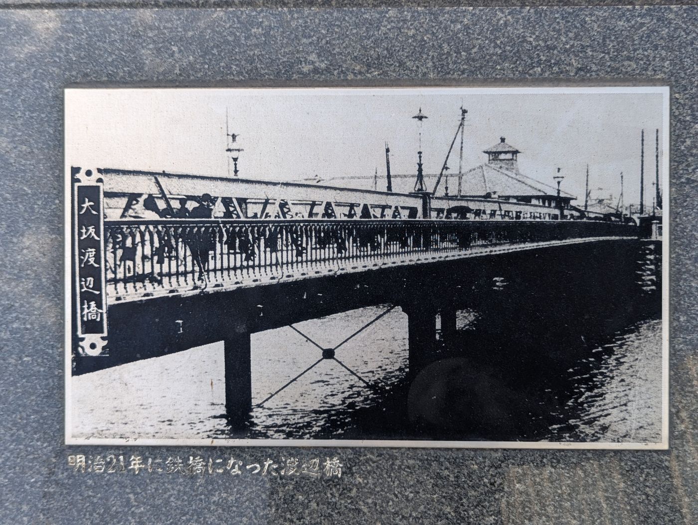 明治21年に鉄橋になった渡辺橋の古写真。大坂渡辺橋と縦書きされた銘板が左側にあり、装飾的な鉄製の欄干を持つアーチ橋の上を市電と人々が行き交っている様子が写されている